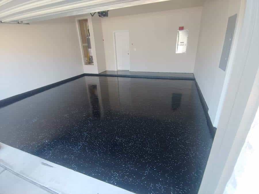 The image shows a room with a glossy black epoxy floor speckled with white and gray flakes, which creates a granite-like appearance. The walls of the room are white, and there's an open garage door indicating that this could be a residential garage space. There's a fire extinguisher on the wall and a door at the back, possibly leading to another room or storage area. The high-gloss finish of the floor is reflective, giving the space a clean and finished look, often desired for modern and functional spaces such as garages or commercial areas.