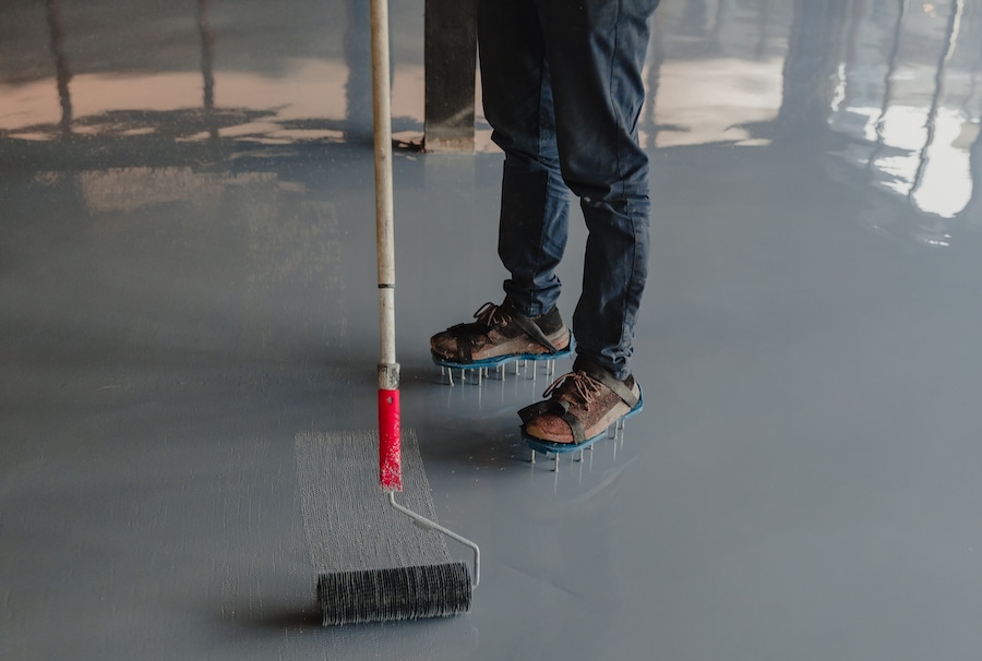The image features a person applying a gray epoxy coating to a concrete floor using a large roller. The individual is wearing spiked shoes, specifically designed for walking on the wet epoxy without leaving marks, and casual attire suggesting hands-on work. The environment reflects light, indicating the glossy nature of the epoxy at this stage. This process is typically part of finishing a floor in spaces such as garages, warehouses, or commercial buildings, where durable and easy-to-clean surfaces are required.