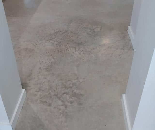 The image depicts a corridor with a decorative concrete floor that has a nuanced, mottled texture, resembling a natural stone or marble finish. The walls are painted white, and the corridor seems to be part of a modern residential or commercial building. The lighting in the hallway reflects softly off the floor's polished surface, indicating a high-quality finish. The seamless transition from the floor to the walls gives the space a sleek and contemporary look.