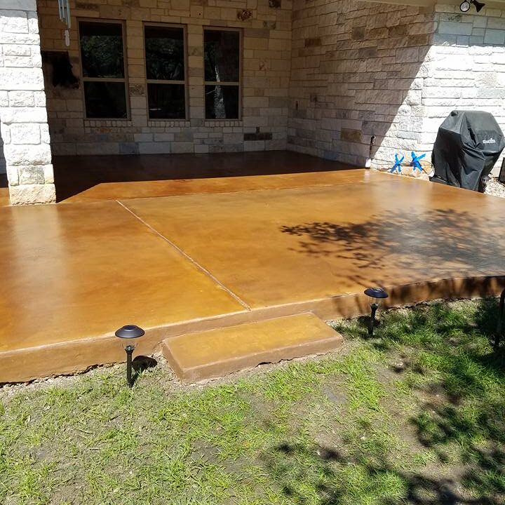 The image showcases a home's outdoor patio area with a newly finished and sealed concrete floor. The concrete has a warm, caramel-like hue that provides a smooth and inviting surface, contrasting pleasantly with the natural stone walls of the house. A couple of small, dark garden lights are installed at the edge of the concrete, highlighting the transition between the lush green grass of the backyard and the patio. The covered patio area promises a comfortable outdoor living space, complete with shade and protection from the elements.