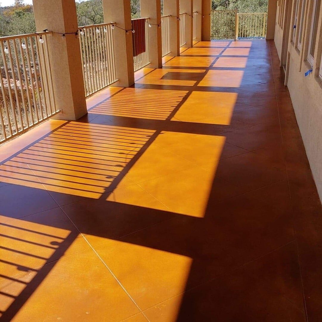 The image showcases a wide corridor with a warm-toned, stained concrete floor that exudes a terracotta hue. The corridor is flanked by a series of columns on one side, casting linear shadows across the floor due to the sunlight, which adds a dynamic, geometric pattern to the scene. The balustrade visible on the far side suggests that the corridor may overlook a lower area or a natural view. The overall ambiance is warm and inviting, highlighted by the rich color of the floor and the interplay of light and shadow.
