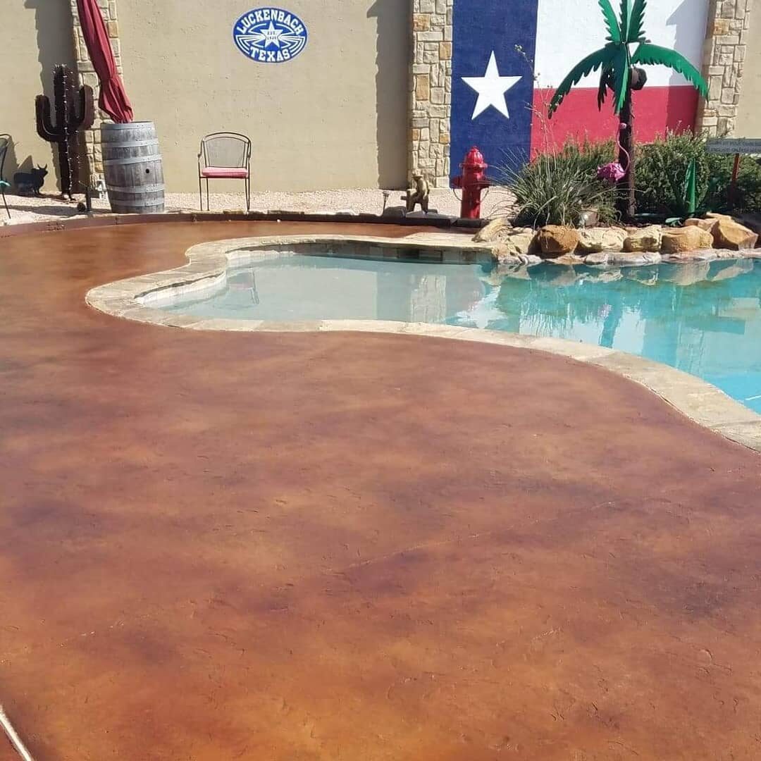 The image shows a residential outdoor area with a freshly coated brown concrete pool deck. The pool has a curved edge, and the area is adorned with a Texas star decoration on the wall, alongside rustic decor elements and landscaping.