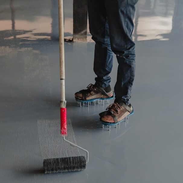 The image features a person applying a gray epoxy coating to a concrete floor using a large roller. The individual is wearing spiked shoes, specifically designed for walking on the wet epoxy without leaving marks, and casual attire suggesting hands-on work. The environment reflects light, indicating the glossy nature of the epoxy at this stage. This process is typically part of finishing a floor in spaces such as garages, warehouses, or commercial buildings, where durable and easy-to-clean surfaces are required.