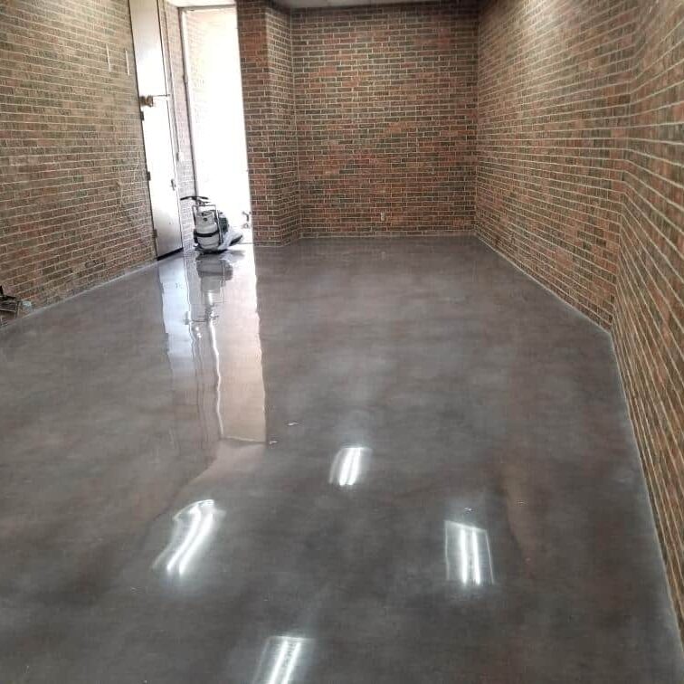 The image displays an interior space with a polished concrete floor that reflects the light, enhancing the room's industrial aesthetic. The floor has a smooth, glossy finish with a dark gray tone. Exposed brick walls on all sides add a rustic charm, indicating this may be a renovated or repurposed building. A white door is open in the distance, and what appears to be a vacuum cleaner or floor polisher can be seen near the entrance, suggesting recent cleaning or maintenance.
