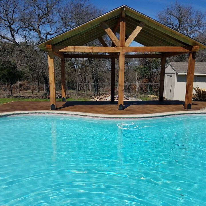 A serene backyard featuring a sparkling, curved-edged swimming pool with clear blue water. In the background, a newly constructed wooden pergola stands on a deck, offering a shaded area for relaxation. The pergola has a symmetrical A-frame roof with sturdy posts. A small shed is visible in the distance, and the area is surrounded by a wire fence with trees beyond, indicating a spacious, private outdoor setting.